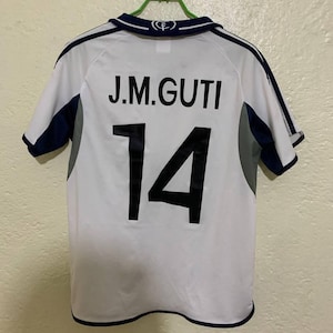 Könnte beinhalten: Ein weißes kurzärmeliges Sporttrikot mit dunkelblauen und grauen Akzenten an den Schultern und am Kragen. Auf der Rückseite steht der Name J.M.GUTI in schwarzer Schrift über der großen schwarzen Nummer 14. Ein kleines blaues Logo ist am Kragen.