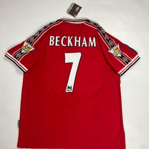 May include: A red Umbro football shirt featuring the name BECKHAM and the number 7 in white on the back. The sleeves have black and white patterned trim with Premier League champion patches. A white collar with black trim and a brand tag are visible.