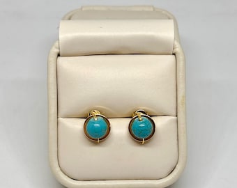 May include: A pair of gold-colored stud earrings with round, turquoise-colored stones. The earrings are displayed in a cream-colored jewelry box with a soft, padded interior. The earrings have a simple, elegant design.