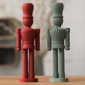 May include: Two decorative wooden soldier figurines. One is solid red with a textured finish, the other is green with vertical striped detailing. Both have tall hats, and stand on a wooden surface.