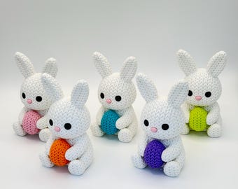 Op de afbeelding: Vijf witte gebreide konijnenbeeldjes, elk met een kleurrijk paasei. De konijnen hebben zwarte ogen en roze neuzen. De eieren zijn roze, oranje, blauw, paars en groen.