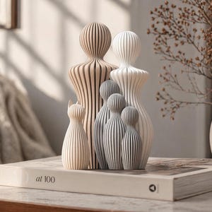 May include: A collection of wooden figurines in varying sizes and colors, arranged on a book. The figurines include two tall, light-colored figures, several smaller gray figures, and a cat-shaped figurine. The book has the text "at 100" on the spine.