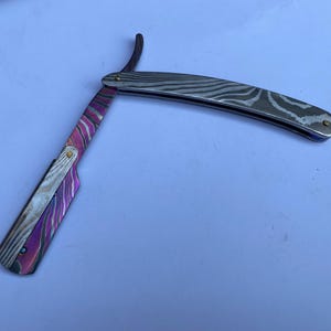 Op de afbeelding: Een recht scheermes met een lemmet en handvat van damaststaal. Het lemmet heeft een zilver en zwart golfpatroon, terwijl het handvat een levendig paars en roze kleurenschema heeft. Het scheermes is open en toont zijn scherpe rand. Lemmetlengte ca. 15 cm.