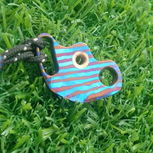 May include: A multi-colored, striped metal keychain with a circular hole and a smaller hole. The keychain is attached to a black and white cord with a matching multi-colored ball. The item is set against a backdrop of green grass.