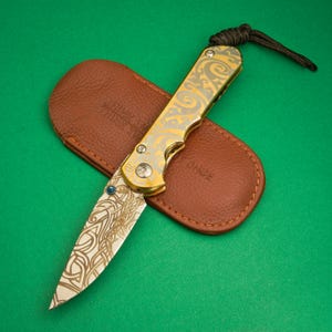 Könnte beinhalten: Ein gold-silbernes Klappmesser mit einem komplizierten Muster auf Klinge und Griff, das auf einer braunen Lederscheide liegt. Das Messer hat einen blauen Edelstein auf der Klinge und eine schwarze Kordel am Griff. Auf der Scheide steht das Wort "ONCE".
