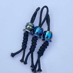 May include: Three decorative lanyard beads with black braided cords. The beads have a metallic finish with iridescent blue, gold, and purple hues. The cords are knotted below the beads and have a loop at the top.
