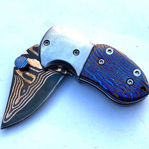 May include: A folding pocket knife with a stainless steel blade and handle. The handle features a blue and bronze striped pattern. The blade is partially open, revealing a copper and black patterned design. The knife has a small blue and bronze thumb stud.