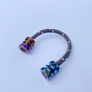 Op de afbeelding: Een klein, U-vormig fidget-speeltje. Het heeft twee metalen, cilindrische uiteinden die zijn verbonden door een grijs en wit gestippeld koord. Eén uiteinde is oranje en paars, de andere is blauw en paars.