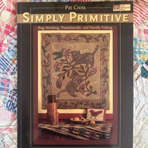 May include: A book titled "Simply Primitive" by Pat Cross, featuring rug hooking, punchneedle, and needle felting. The cover displays a framed image of two cats among floral elements. The book is set against a colorful patchwork quilt.