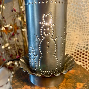 May include: A silver metal candle holder with a snowman design. The cylindrical holder has a series of punched-out holes forming the snowman and other decorative patterns. The candle holder sits on a scalloped base.