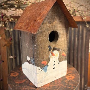 May include: A rustic wooden birdhouse with a rusty metal roof. The birdhouse is painted with a snowman and birds on a snowy landscape. The birdhouse is sitting on a decorative, round, wooden stand.