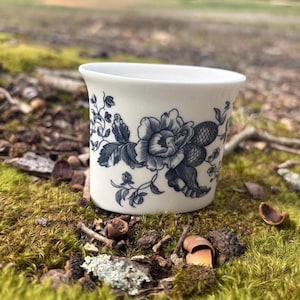 May include: A white ceramic cup with a flared rim, adorned with a detailed blue floral pattern. The design features large flowers and foliage. The cup is set against a natural, mossy background.