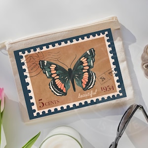 May include: A beige canvas zipper pouch featuring a rectangular graphic of a vintage postage stamp. The stamp has a dark blue scalloped border, a brown background, a butterfly with orange and black wings, and text reading 5 CENTS, beautiful, 1954.