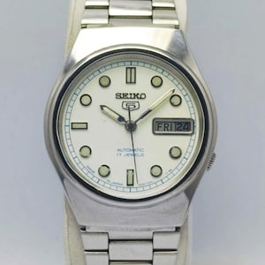 Puede incluir: Un reloj de pulsera automático Seiko 5 plateado con esfera blanca. El reloj presenta una pantalla de día y fecha, agujas e indicadores luminosos, y una correa de acero inoxidable. La esfera del reloj muestra el texto "SEIKO 5 AUTOMATIC 17 JEWELS".