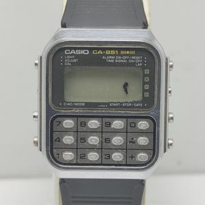May include: A vintage Casio CA-851 digital watch with a calculator keypad. The watch has a silver-tone case, a black rubber strap, and a digital display. The face includes buttons for light, adjust, and cal. The watch also has a calculator function.