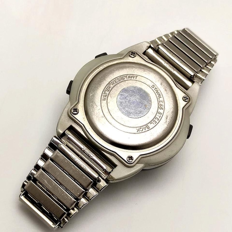 Puede incluir: Un reloj digital plateado con una correa de acero inoxidable, visto desde atr&aacute;s. La esfera del reloj es redonda y tiene las palabras "Water Resistant" y "Stainless Steel Back" impresas. La correa est&aacute; hecha de eslabones met&aacute;licos.