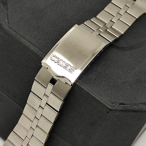 May include: A close-up of a silver-toned Seiko watch band. The band is made of rectangular links and features a clasp with the Seiko logo. The watch band is on a dark surface.