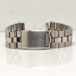 May include: A silver-toned Seiko watch band with a brushed metal finish. The band is composed of rectangular links and features a fold-over clasp with the Seiko logo. The band is designed to be attached to a watch.