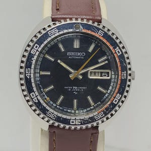 Puede incluir: Reloj automático Seiko vintage con esfera azul oscuro, bisel giratorio con marcas blancas y naranjas, y correa de cuero marrón. La esfera del reloj muestra el día y la fecha.