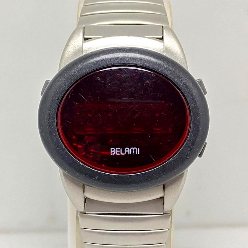 Puede incluir: Un reloj digital Belami vintage con una correa de metal plateado. La esfera del reloj es de forma ovalada roja oscura, rodeada por un bisel negro. La palabra "BELAMI" est&aacute; impresa en blanco en la esfera.