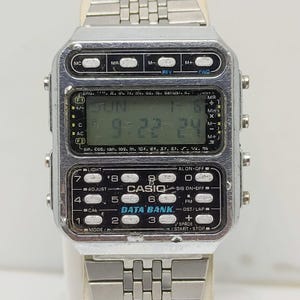 Może przedstawiać: Zegarek cyfrowy Casio Data Bank w stylu vintage ze srebrnym metalowym paskiem i kopertą. Tarcza zegarka wyświetla godzinę, dzień i datę. Posiada wiele przycisków i funkcji, w tym kalkulator i przechowywanie danych.