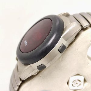Puede incluir: Primer plano de un reloj digital plateado con un bisel gris oscuro y una pantalla roja. El reloj tiene una correa plateada a juego y dos botones grises en el lateral. La esfera del reloj muestra la palabra "LIFE".