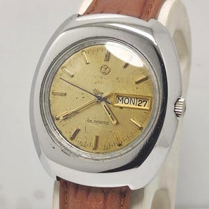 May include: A vintage Oldmatic wristwatch with a gold-toned face and a silver bezel. The watch shows the day and date, with a brown leather strap. The watch face has gold hands and markers. The watch is displayed on a white surface.