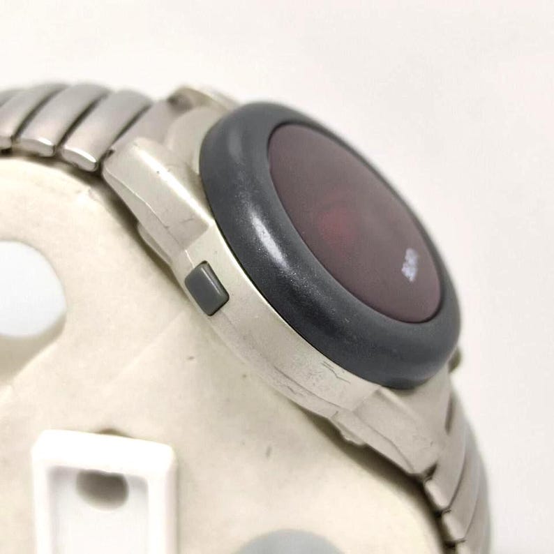 Puede incluir: Un reloj digital plateado con un bisel gris oscuro y una pantalla roja. El reloj tiene una correa de metal segmentada y un peque&ntilde;o bot&oacute;n gris en el lateral. El reloj est&aacute; en &aacute;ngulo, mostrando el lateral y la esfera.