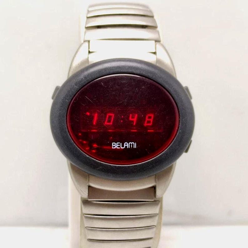 Puede incluir: Un reloj de pulsera digital vintage con una correa plateada y un bisel negro. La esfera del reloj muestra la hora, 10:48, en n&uacute;meros digitales rojos. La marca "BELAMI" est&aacute; impresa debajo de la pantalla de la hora.