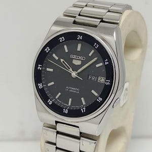 May include: A silver Seiko 5 automatic wristwatch with a black dial. The watch features a day and date display, luminous hands, and a stainless steel band. The dial displays the text "SEIKO", "AUTOMATIC", and "17 JEWELS".