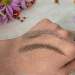 Peut inclure: Une main présentant deux exemples de sourcils. L'un est un sourcil dessiné, d'apparence naturelle, et l'autre est un pochoir. La peau est claire, et les sourcils sont bruns moyens. Le fond comprend des fleurs.