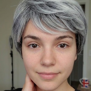 May include: A person with fair skin, freckles, and brown eyes, wearing a black shirt, and a silver nose ring. They have short, layered, gray and white hair. A mug with a Union Jack design is visible in the background.