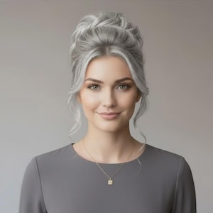May include: A woman with silver hair styled in an updo, wearing a gray dress and a gold necklace with a square pendant. The background is a solid gray. The woman is smiling.