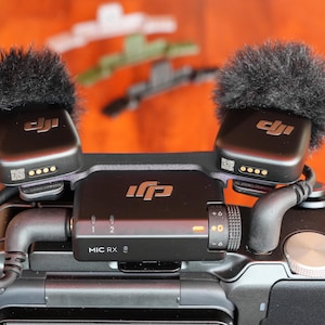 Op de afbeelding: Een zwart DJI draadloos microfoonsysteem gemonteerd op een camera. De set bevat een ontvanger met het label MIC RX en een draaiknop, en twee zenders met zwarte harige windschermen. De ontvanger is verbonden met de camera via een zwarte kabel.
