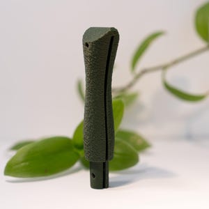 Peut inclure: Une poignée vert olive foncé avec une finition mate et texturée. La poignée présente une fente verticale le long du corps principal et une base cylindrique plus petite et lisse avec un trou visible près du bas pour la fixation.