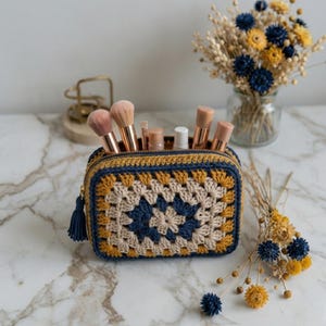 May include: A crochet makeup bag in a granny square design. The bag is navy blue, tan, and mustard yellow, with a navy tassel on the zip. The bag contains makeup brushes and cosmetic products. The bag is handmade.
