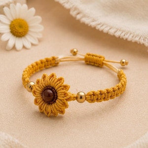 May include: Yellow macrame bracelet featuring a sunflower design with a brown center and gold beads. The adjustable bracelet is made of woven yellow thread. A daisy flower is in the background.