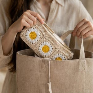 以下が含まれることがあります： ベージュのかぎ針編みのペンケース。デイジーの花のデザインで、お揃いのベージュのトートバッグに入っています。ペンケースはファスナー開閉式で、持ち手が付いています。デイジーの花は白い花びらと黄色の中心部があります。