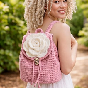 May include: A pink crochet backpack with a large white flower applique on the front flap. The backpack has adjustable straps and a drawstring closure. The bag is made of a soft, textured yarn.