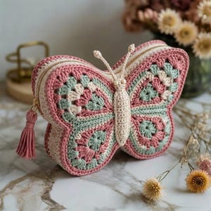 Puede incluir: Bolso hecho a mano con forma de mariposa, tejido a ganchillo en tonos rosa, crema y verde menta. El bolso tiene cierre de cremallera con borla y detalles de ganchillo. Las antenas y el cuerpo de la mariposa también están tejidos.