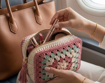 以下が含まれることがあります： ピンク、クリーム、ミントグリーンのグラニースクエア模様の、手編みの化粧ポーチ。ゴールドのジッパーと、お揃いのタッセルが付いています。中には口紅、コンパクト、マスカラが入っています。背景には茶色のレザートートバッグがあります。
