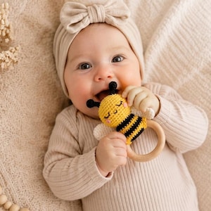 May include: A baby wearing a beige headband and long-sleeved onesie smiles while holding a yellow and black bee-shaped wooden teether. The baby is lying on a beige blanket, with decorative wooden beads and dried flowers nearby.