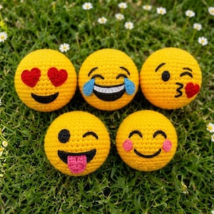 May include: Five round, yellow, crocheted emoji balls featuring different facial expressions: heart eyes, laughing with tears, blowing a kiss with a heart, winking with a tongue out, and a smiling face with pink cheeks, all arranged in a group.