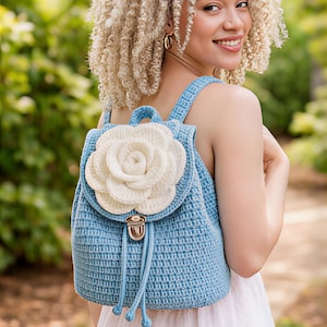 Peut inclure: Un sac à dos en crochet bleu clair avec une grande fleur blanche appliquée sur le devant. Le sac à dos a des bretelles réglables et une fermeture à cordon. Le sac est fait d'un fil texturé et possède un fermoir en métal. L'arrière-plan est une scène extérieure floue.