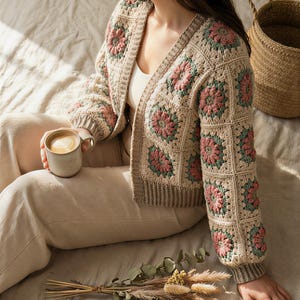 May include: A cream-coloured crochet cardigan featuring a floral design in pink and green. The cardigan has an open front and long sleeves. A person is holding a mug of coffee. A woven basket and a bouquet of dried flowers are also present.
