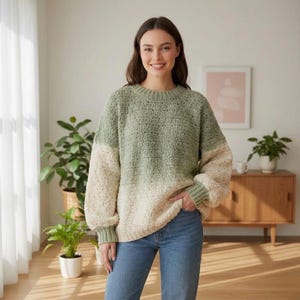 Puede incluir: Un suéter de crochet de dos tonos en verde salvia y crema. El suéter tiene cuello redondo, mangas largas y puños y dobladillo acanalados. La modelo lleva vaqueros azules. El suéter es una prenda cómoda y acogedora.