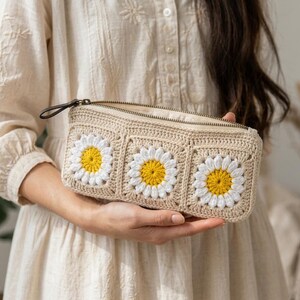 Floral crochet toiletry pouch with daisy granny square design, structured rectangular shape, zipper closure, and neutral cotton yarn aesthetic for beauty essentials.