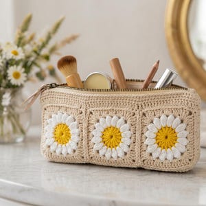 Pode incluir: Uma bolsa de cosméticos de crochê bege com três desenhos de flores de margarida. A bolsa está aberta, revelando pincéis e tubos de maquiagem. A bolsa tem fecho de zíper e está sobre uma superfície branca.