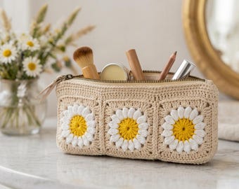 以下が含まれることがあります： ベージュのかぎ針編みの化粧ポーチ。デイジーの花のデザインが3つあります。バッグは開いており、メイクブラシやチューブが見えます。バッグはジッパーで開閉し、白い表面に置かれています。