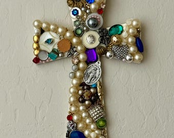 May include: A decorative cross adorned with faux pearls, colorful gemstones, and various embellishments. The cross features a mix of blue, green, red, and gold accents, creating a unique and ornate design. The cross is a religious item.